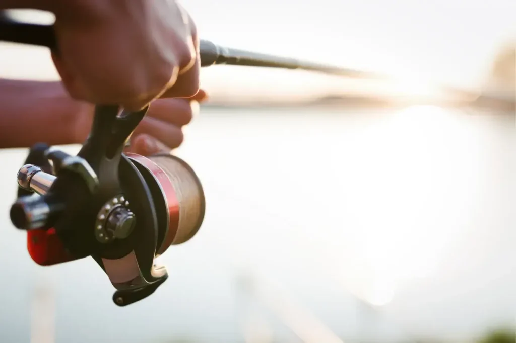 A close-up of an angler holding a spinning reel while casting at sunrise, with a lake in the background.