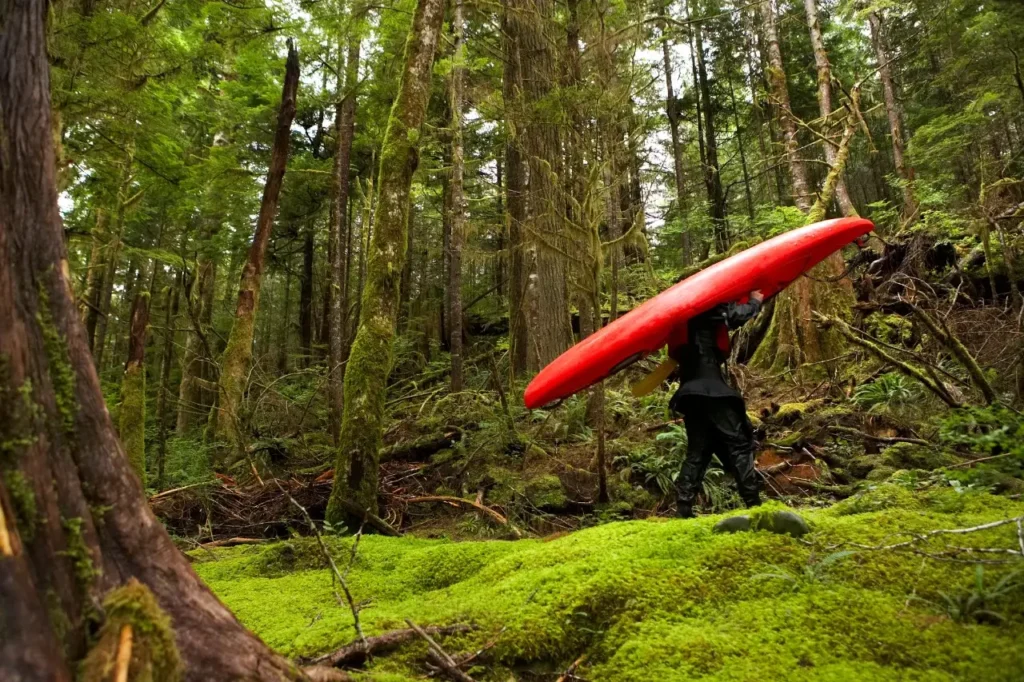 An angler carries a lightweight fishing kayak on their shoulder while walking through a forest, highlighting its portability and ease of transport.