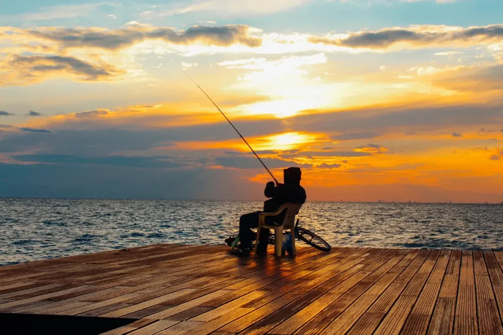 An angler enjoys a peaceful fishing session on a wooden dock at sunset, casting a line into the calm sea under a colorful evening sky.