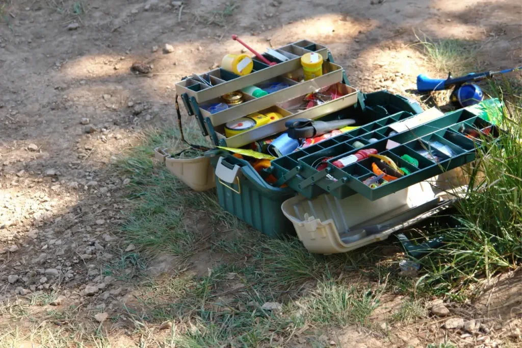An open fishing tackle box filled with essential fishing items, including lures, hooks, fishing line, and tools, is placed on the ground near a fishing spot.